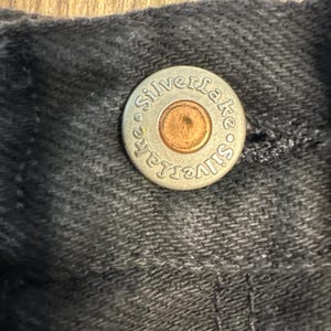 K&ouml;nnte beinhalten: Nahaufnahme eines silbernen Knopfes mit dem gepr&auml;gten Text "Silverlake". Der Knopf ist an einem dunkelgrauen Stoff befestigt.