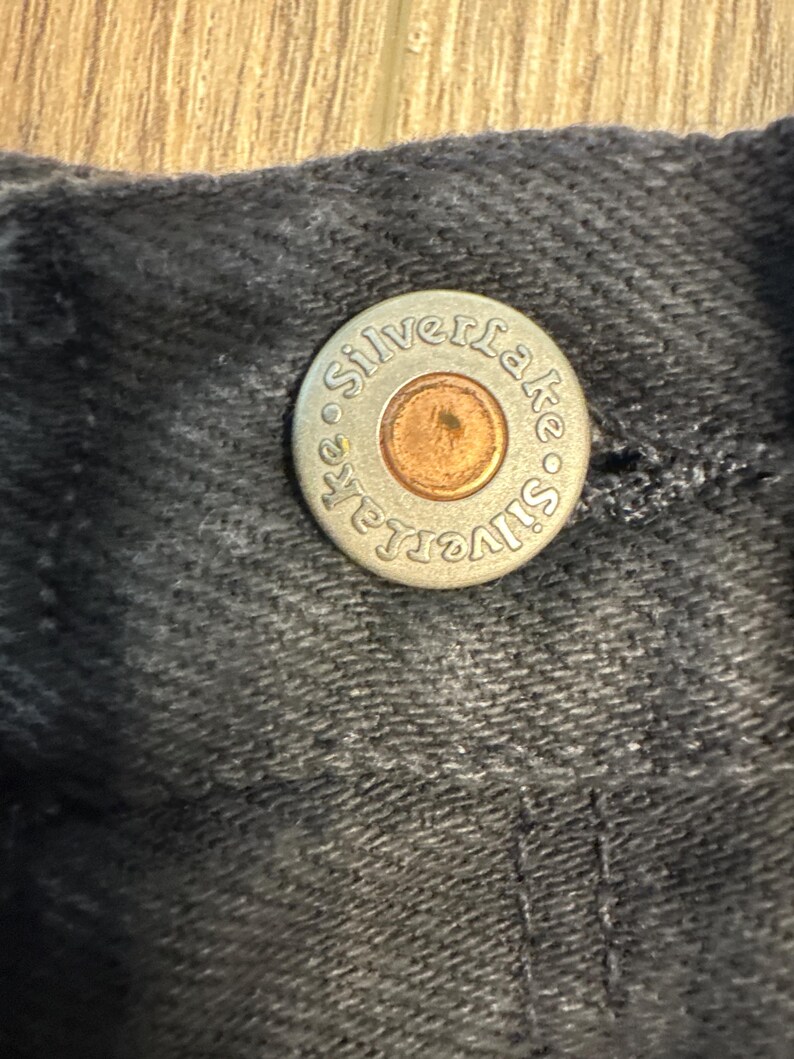 K&ouml;nnte beinhalten: Nahaufnahme eines silbernen Knopfes mit dem gepr&auml;gten Text "Silverlake". Der Knopf ist an einem dunkelgrauen Stoff befestigt.