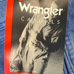 May include: A close-up of a Wrangler Casuals tag attached to blue denim fabric. The tag is black and white with a grayscale image of a person in denim overalls. The tag is printed in the U.S.A.