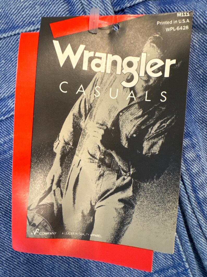 May include: A close-up of a Wrangler Casuals tag attached to blue denim fabric. The tag is black and white with a grayscale image of a person in denim overalls. The tag is printed in the U.S.A.