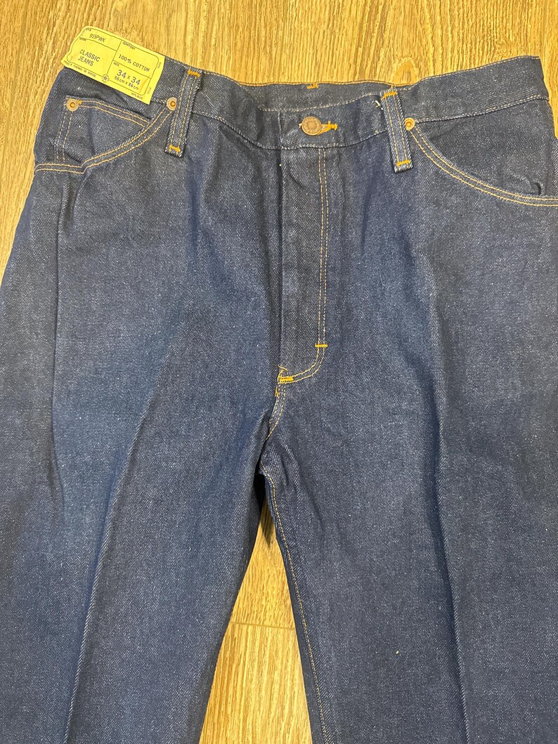 May include: Dark blue classic jeans with a straight leg cut. The jeans have a button and zipper closure, front and back pockets, and yellow stitching. A tag reads "CLASSIC JEANS" and "34x34".