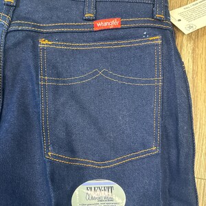 May include: Close-up of dark blue Wrangler jeans with a back pocket and a red Wrangler label. The jeans have orange stitching and a Flex-Fit Comfort Wear label. The jeans have a stretch waistband.