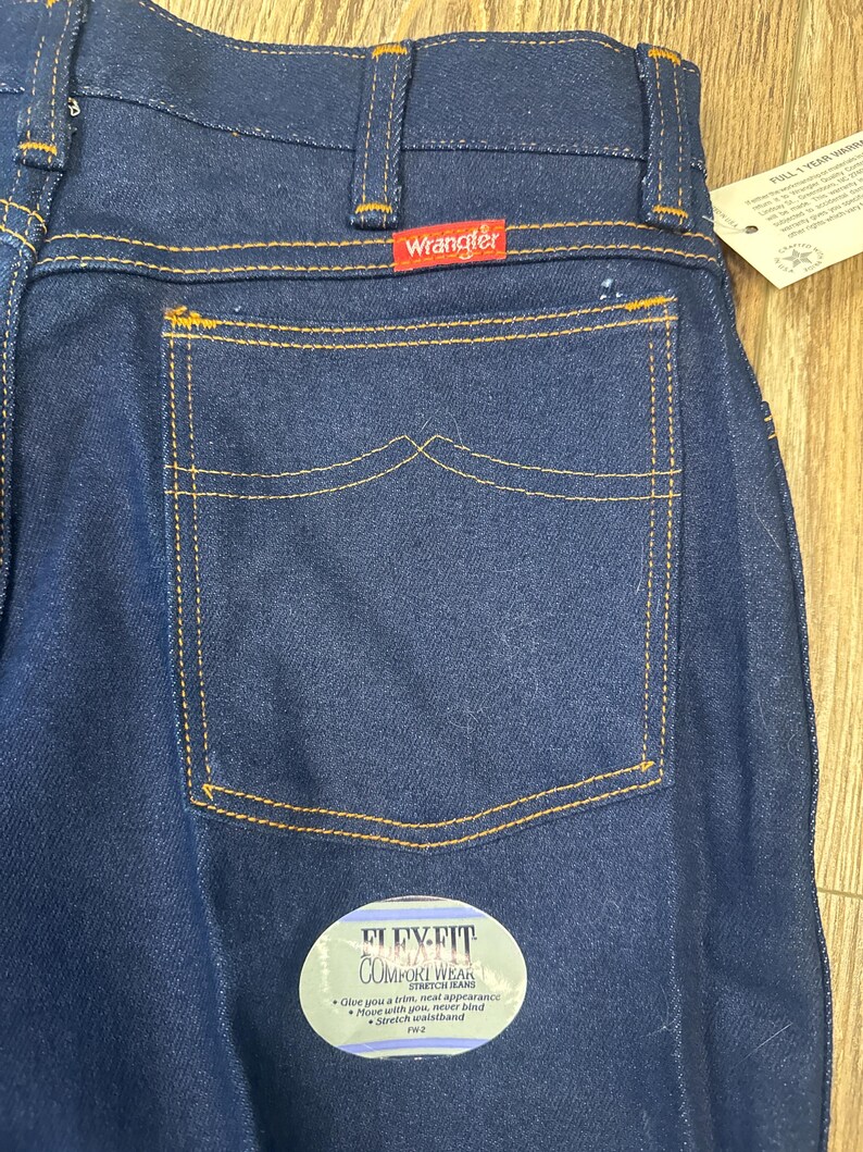 May include: Close-up of dark blue Wrangler jeans with a back pocket and a red Wrangler label. The jeans have orange stitching and a Flex-Fit Comfort Wear label. The jeans have a stretch waistband.