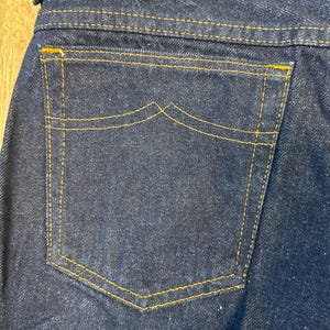 May include: Close-up of the back pocket of dark blue denim jeans. The pocket is outlined with yellow stitching, and the top of the pocket has a unique curved design. The jeans are made of a sturdy denim material.