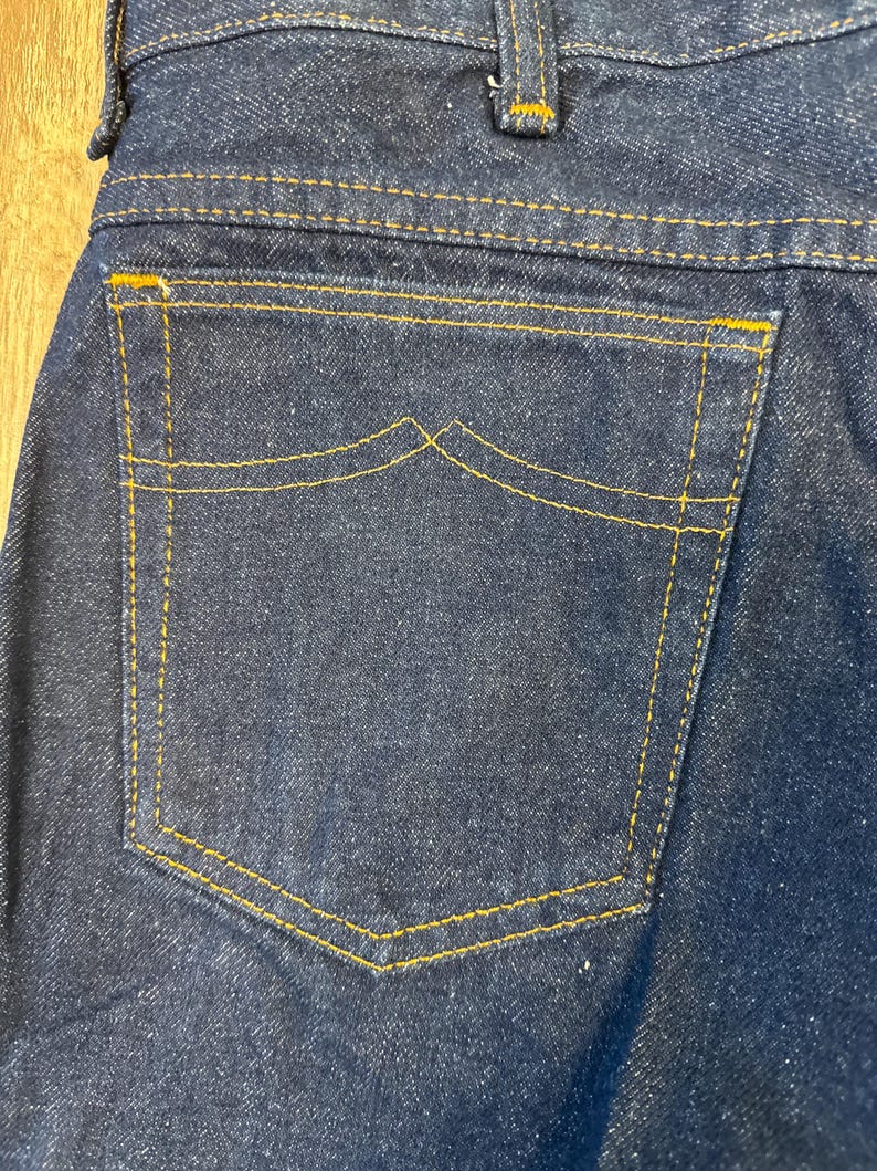 May include: Close-up of the back pocket of dark blue denim jeans. The pocket is outlined with yellow stitching, and the top of the pocket has a unique curved design. The jeans are made of a sturdy denim material.