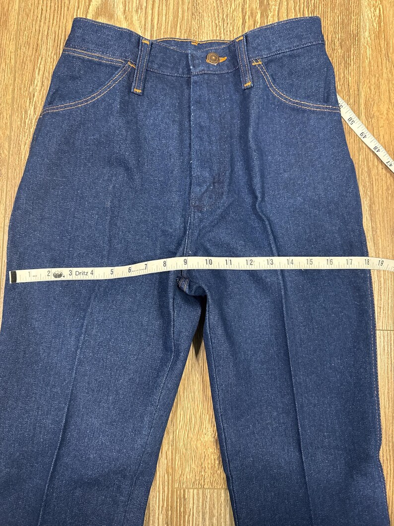 K&ouml;nnte beinhalten: Nahaufnahme einer blauen Jeans mit Knopfverschluss und Taschen. Ein Ma&szlig;band zeigt die Bundweite. Die Jeans hat ein klassisches, gerades Bein-Design.