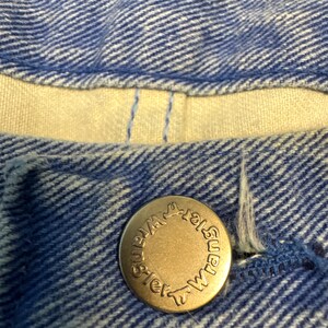 May include: Close-up of blue denim fabric with a brass button. The button is embossed with the word "Wrangler" in a circular pattern. The denim has a textured appearance.