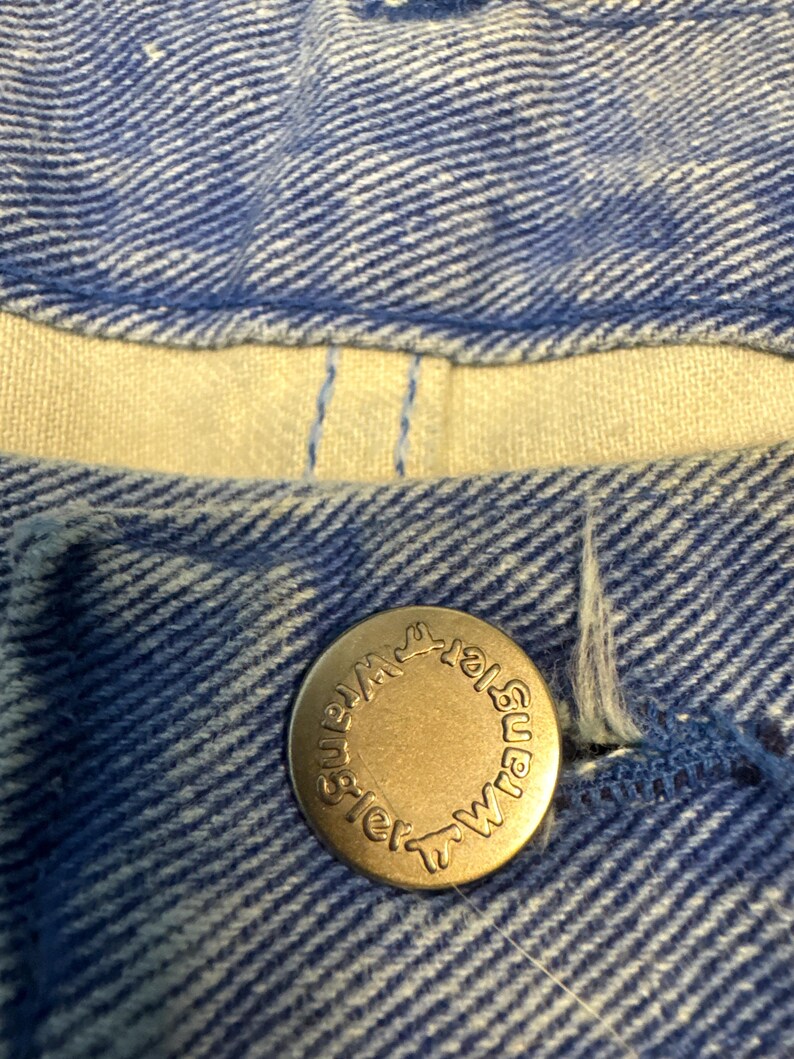 May include: Close-up of blue denim fabric with a brass button. The button is embossed with the word "Wrangler" in a circular pattern. The denim has a textured appearance.