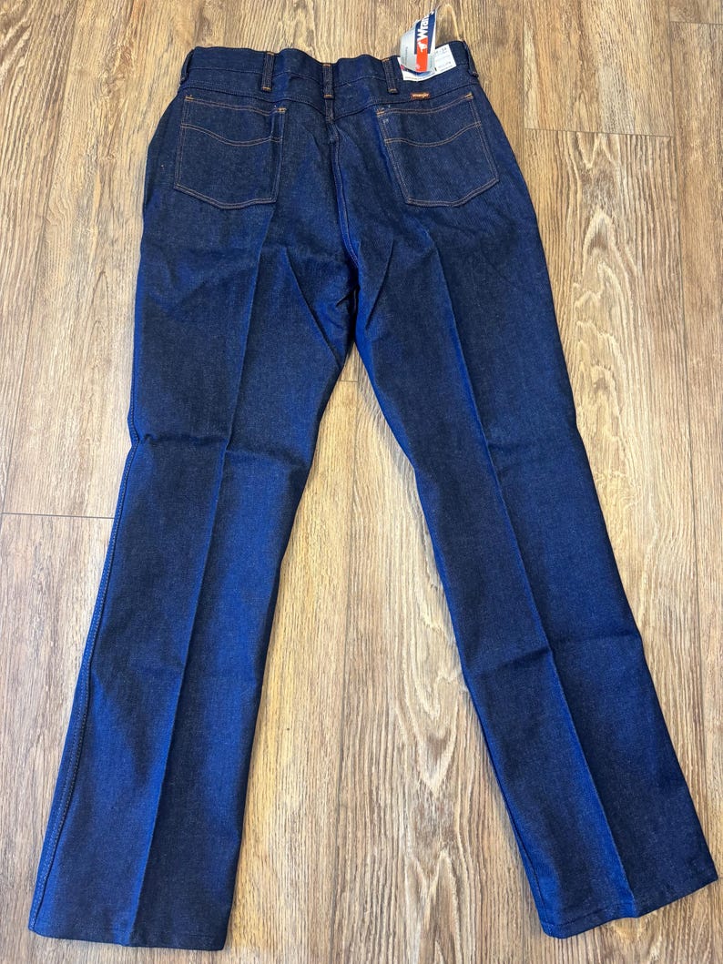 May include: Dark blue denim jeans with a classic five-pocket design. The jeans have a straight leg cut and feature a Wrangler tag. The back pockets have a stitched design. The jeans are made of a durable denim material.