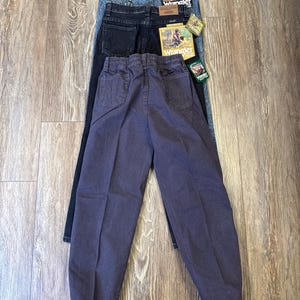 Może przedstawiać: Trzy pary dżins&oacute;w Wrangler w r&oacute;żnych odcieniach: acid wash, ciemnoniebieski i wyblakły fiolet. Dżinsy leżą na drewnianej powierzchni, prezentując swoje r&oacute;żne style i kolory. Każda para ma naszywkę Wrangler.