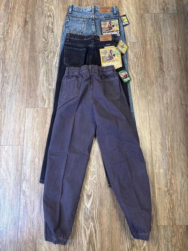 Może przedstawiać: Trzy pary dżins&oacute;w Wrangler w r&oacute;żnych odcieniach: acid wash, ciemnoniebieski i wyblakły fiolet. Dżinsy leżą na drewnianej powierzchni, prezentując swoje r&oacute;żne style i kolory. Każda para ma naszywkę Wrangler.
