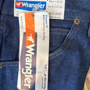 May include: A blue denim pair of Wrangler jeans with a white tag attached. The tag says "Full 1 Year Warranty", "Wrangler Brand", "34 x 32", "86cm x 81cm", "100% Cotton", "85311PW", "Proven Fit with Lasting Quality", "Made in U.S.A."