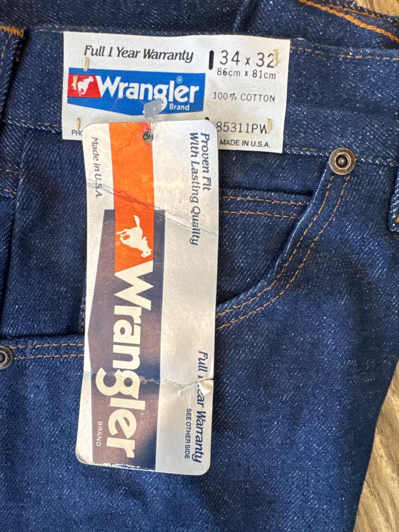 May include: A blue denim pair of Wrangler jeans with a white tag attached. The tag says "Full 1 Year Warranty", "Wrangler Brand", "34 x 32", "86cm x 81cm", "100% Cotton", "85311PW", "Proven Fit with Lasting Quality", "Made in U.S.A."