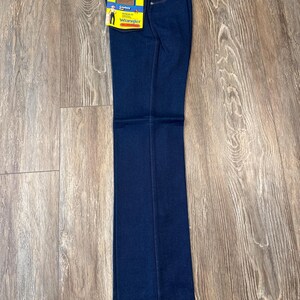 May include: Dark blue Wrangler jeans with a straight leg. The jeans have a tag attached with the Wrangler logo and a cowboy graphic. The jeans are folded and laid flat on a wooden surface.