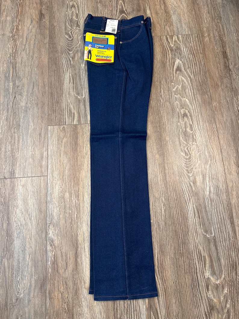 May include: Dark blue Wrangler jeans with a straight leg. The jeans have a tag attached with the Wrangler logo and a cowboy graphic. The jeans are folded and laid flat on a wooden surface.