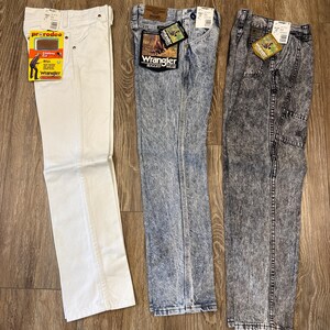 K&ouml;nnte beinhalten: Drei Jeanshosen von Wrangler werden auf einer Holzoberfl&auml;che gezeigt. Die Jeans sind wei&szlig;, blau mit S&auml;urewaschung und grau mit S&auml;urewaschung. Jede Hose hat Etiketten. Die wei&szlig;e Jeans hat ein "ProRodeo"-Etikett. Die blauen und grauen Jeans haben "Wrangler Rugged Wear"-Etiketten.