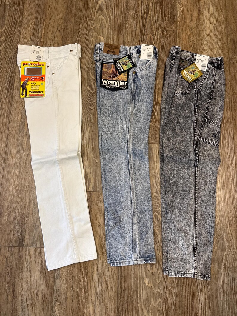 K&ouml;nnte beinhalten: Drei Jeanshosen von Wrangler werden auf einer Holzoberfl&auml;che gezeigt. Die Jeans sind wei&szlig;, blau mit S&auml;urewaschung und grau mit S&auml;urewaschung. Jede Hose hat Etiketten. Die wei&szlig;e Jeans hat ein "ProRodeo"-Etikett. Die blauen und grauen Jeans haben "Wrangler Rugged Wear"-Etiketten.