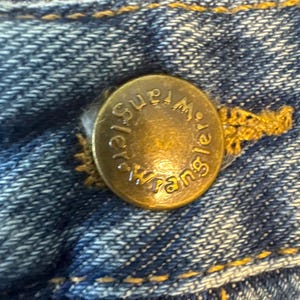 May include: Close-up of a gold-colored metal button with the word "Wrangler" embossed on it. The button is attached to a pair of blue denim jeans.