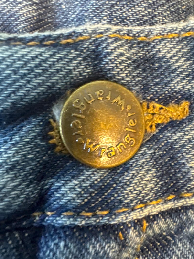 May include: Close-up of a gold-colored metal button with the word "Wrangler" embossed on it. The button is attached to a pair of blue denim jeans.
