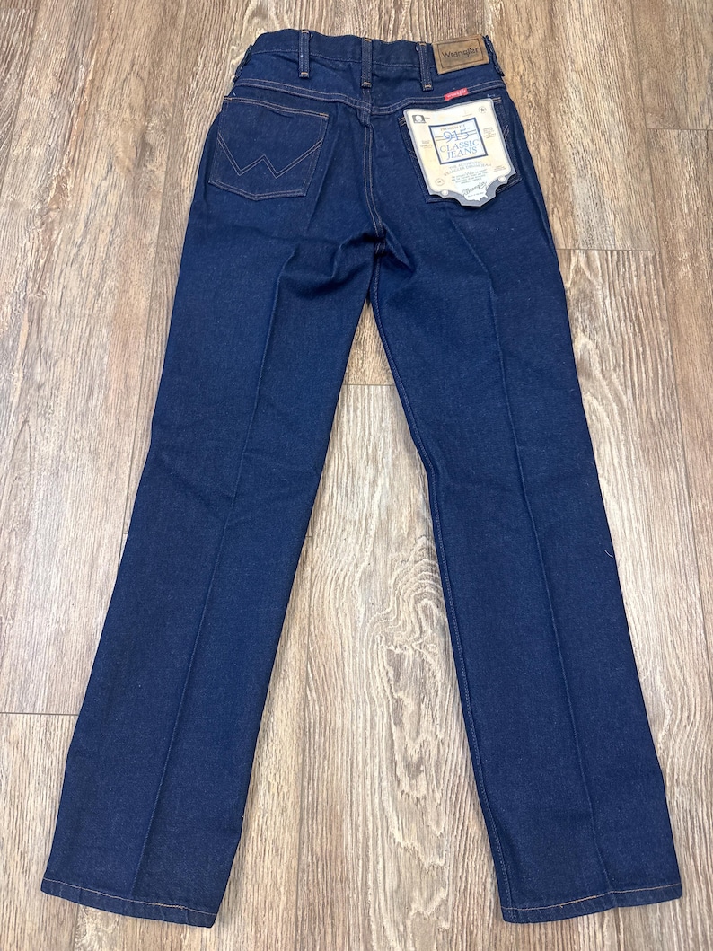 May include: Dark blue denim jeans with a classic fit. The jeans feature the Wrangler logo on the back pocket and a tag with the text "915 Classic Jeans". The jeans have a straight leg and are made of a durable denim material.