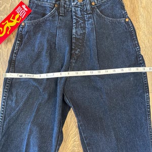 May include: Dark blue denim Wrangler jeans with a high waist and pleats. The jeans have a straight leg and classic five-pocket design. A measuring tape is laid across the waist. A red and yellow RIATA tag is visible.