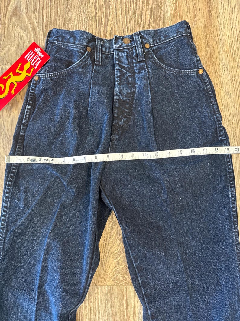 May include: Dark blue denim Wrangler jeans with a high waist and pleats. The jeans have a straight leg and classic five-pocket design. A measuring tape is laid across the waist. A red and yellow RIATA tag is visible.
