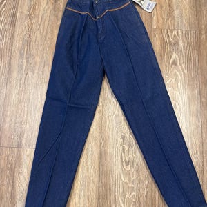 Puede incluir: Un par de pantalones vaqueros de mezclilla azul oscuro con detalles de costura marr&oacute;n alrededor de la cintura. Los pantalones tienen un corte de pierna recta y est&aacute;n hechos por Wrangler.