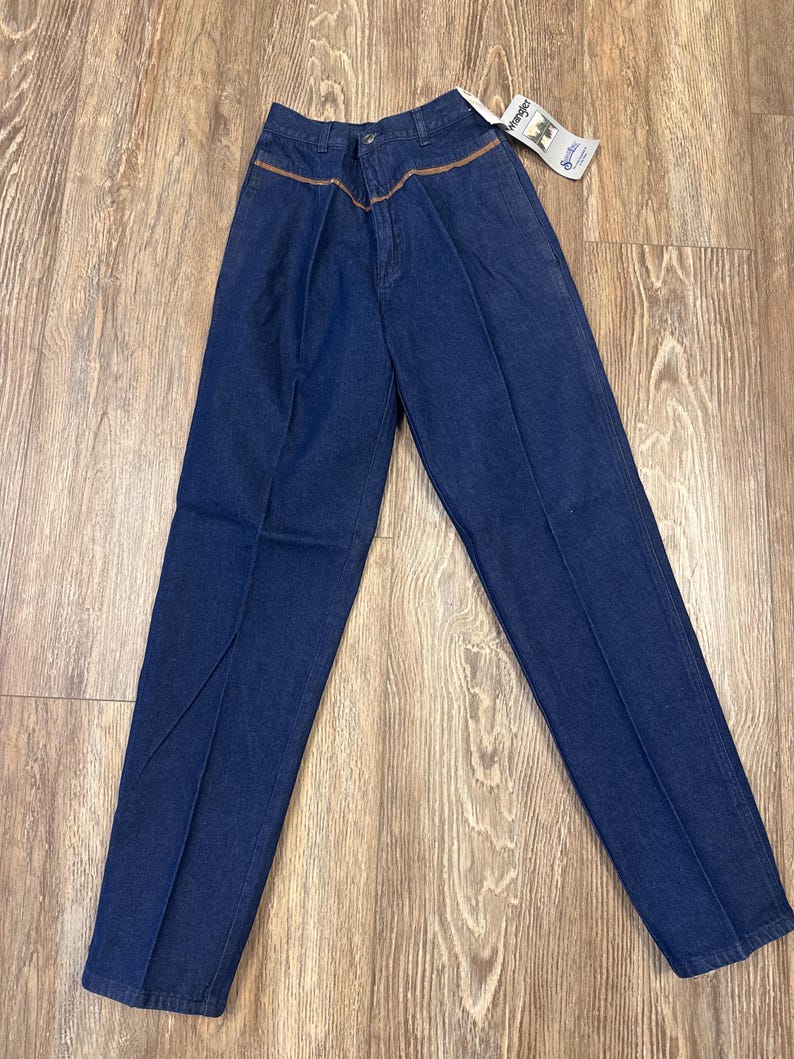 Puede incluir: Un par de pantalones vaqueros de mezclilla azul oscuro con detalles de costura marr&oacute;n alrededor de la cintura. Los pantalones tienen un corte de pierna recta y est&aacute;n hechos por Wrangler.