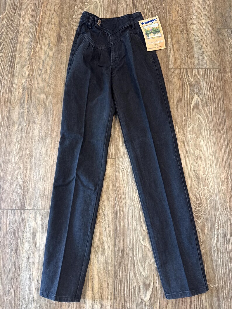 May include: Dark wash denim Wrangler jeans with a high waist and pleated front. The jeans have a button closure and a straight leg. A Wrangler tag is attached to the jeans.