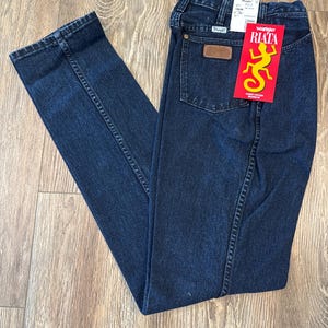 May include: Dark blue Wrangler Riata jeans with a brown leather patch on the back pocket. A red tag with a yellow lizard graphic and the word "RIATA" is attached. The jeans are folded on a wooden surface.