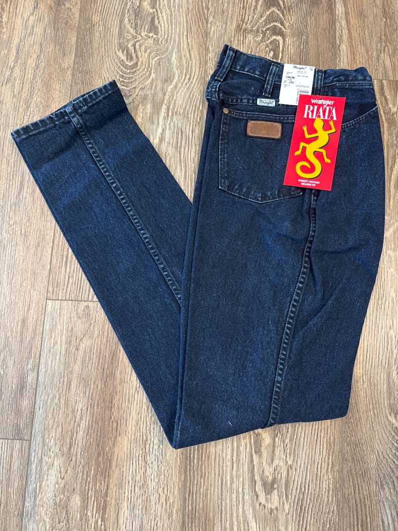 May include: Dark blue Wrangler Riata jeans with a brown leather patch on the back pocket. A red tag with a yellow lizard graphic and the word "RIATA" is attached. The jeans are folded on a wooden surface.