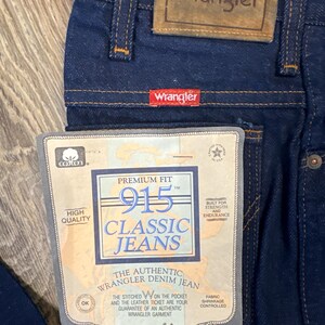 May include: Close-up of dark blue Wrangler 915 Classic Jeans. The jeans have a leather patch and a red Wrangler tag. A paper tag reads "Premium Fit" and "The Authentic Wrangler Denim Jean".