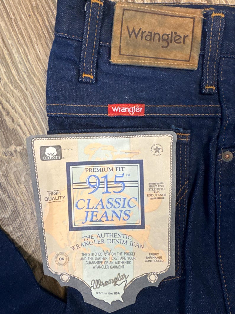 May include: Close-up of dark blue Wrangler 915 Classic Jeans. The jeans have a leather patch and a red Wrangler tag. A paper tag reads "Premium Fit" and "The Authentic Wrangler Denim Jean".