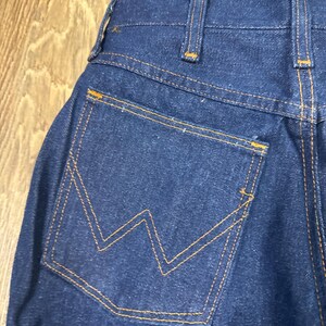 K&ouml;nnte beinhalten: Nahaufnahme der R&uuml;ckseite einer dunkelblauen Jeans. Die Jeans hat eine einzelne Ges&auml;&szlig;tasche mit einem markanten orangefarbenen Nahtdesign. Die N&auml;hte und G&uuml;rtelschlaufen sind ebenfalls mit orangefarbenem Faden akzentuiert, was einen klassischen Look erzeugt.