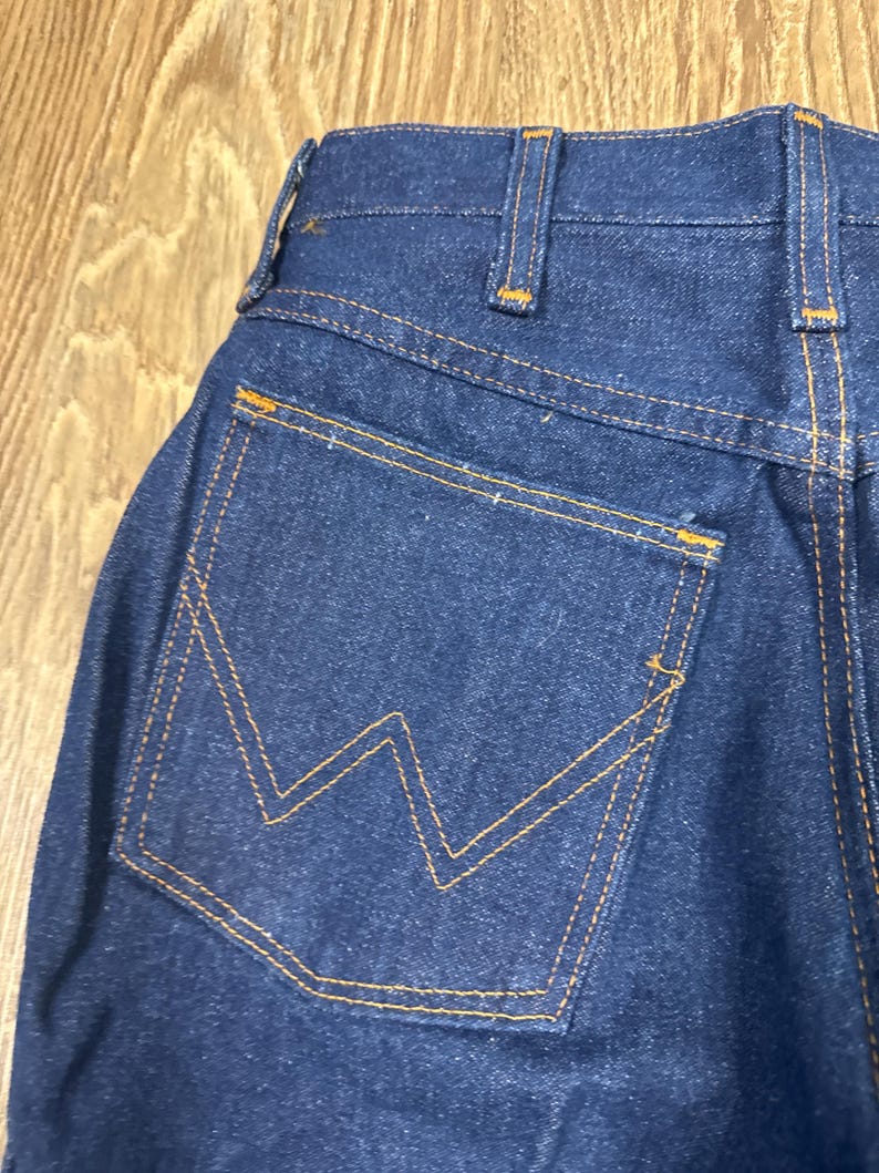 K&ouml;nnte beinhalten: Nahaufnahme der R&uuml;ckseite einer dunkelblauen Jeans. Die Jeans hat eine einzelne Ges&auml;&szlig;tasche mit einem markanten orangefarbenen Nahtdesign. Die N&auml;hte und G&uuml;rtelschlaufen sind ebenfalls mit orangefarbenem Faden akzentuiert, was einen klassischen Look erzeugt.