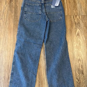 May include: Blue denim jeans with a straight leg cut. The jeans have a metal buckle on the back and a tag that reads "Wrangler Blues". The jeans have two back pockets and a classic five-pocket design.