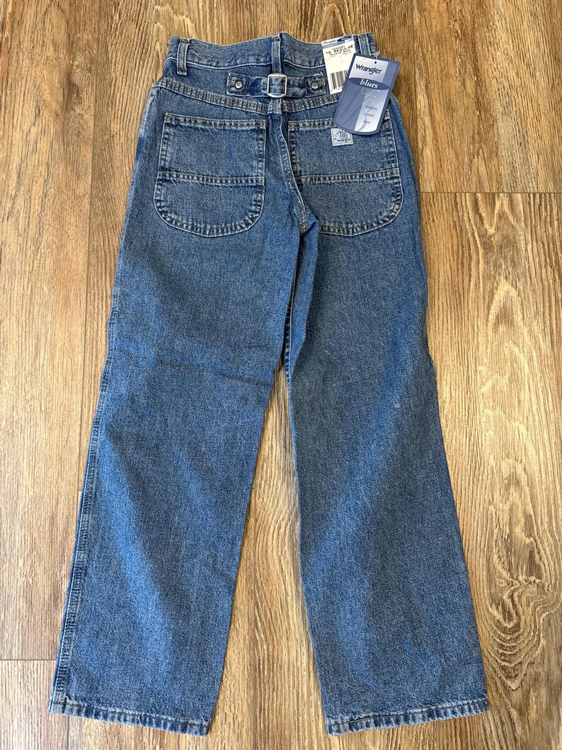 May include: Blue denim jeans with a straight leg cut. The jeans have a metal buckle on the back and a tag that reads "Wrangler Blues". The jeans have two back pockets and a classic five-pocket design.