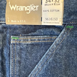 May include: Close-up of blue denim Wrangler jeans. The tag reads "34 x 32" and "Made in U.S.A." The jeans are 34 inches at the waist and 32 inches in length. The fabric is 100% cotton.