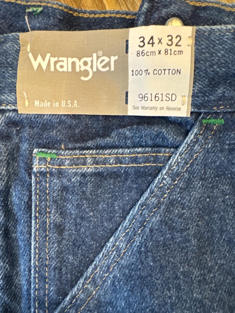 May include: Close-up of blue denim Wrangler jeans. The tag reads "34 x 32" and "Made in U.S.A." The jeans are 34 inches at the waist and 32 inches in length. The fabric is 100% cotton.