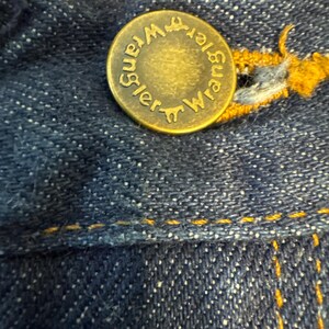 May include: Close-up of a brass button on dark blue denim jeans. The button is embossed with the word "Wrangler" in a circular pattern. Yellow stitching is visible around the button and along the seams of the jeans.