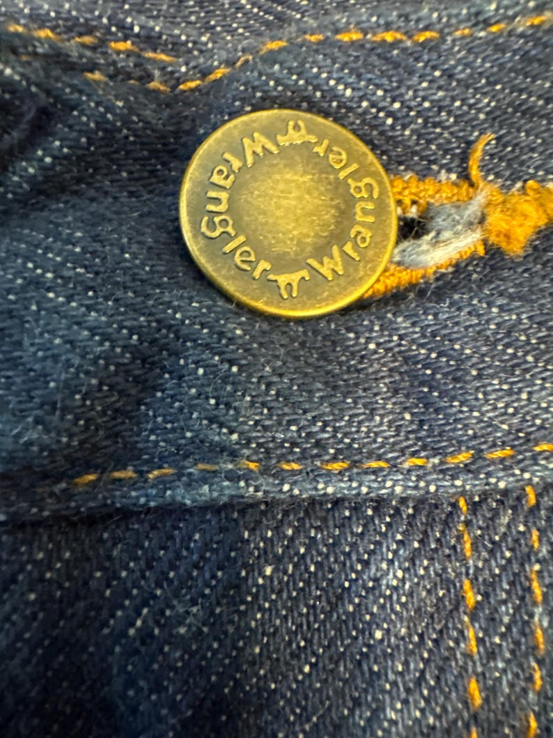 May include: Close-up of a brass button on dark blue denim jeans. The button is embossed with the word "Wrangler" in a circular pattern. Yellow stitching is visible around the button and along the seams of the jeans.