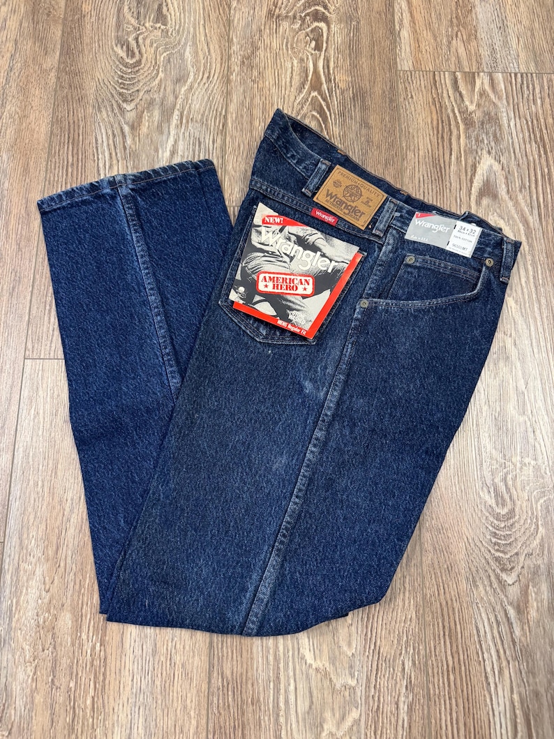 May include: Dark blue Wrangler jeans with a classic five-pocket design. The jeans have a straight leg cut and feature the Wrangler brand label on the back pocket and waistband. A red and white tag reads "American Hero".