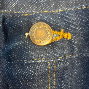 May include: Close-up of dark blue denim fabric with a copper-colored button. The button is embossed with the word "Wrangler" in a circular pattern. Yellow stitching is visible around the button and along the denim seams, highlighting the details of the garment.
