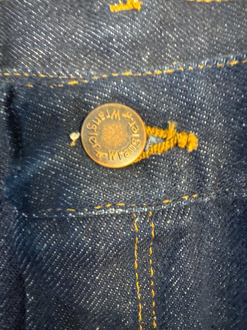 May include: Close-up of dark blue denim fabric with a copper-colored button. The button is embossed with the word "Wrangler" in a circular pattern. Yellow stitching is visible around the button and along the denim seams, highlighting the details of the garment.