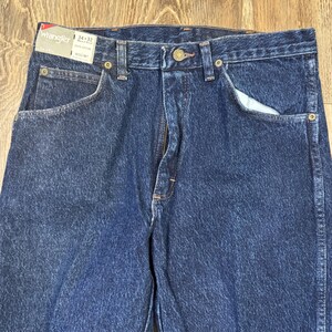May include: A close-up shot of blue denim Wrangler jeans. The jeans feature a classic five-pocket design, a zip fly, and a button closure. The label indicates a 34x32 size. The denim has a medium wash.