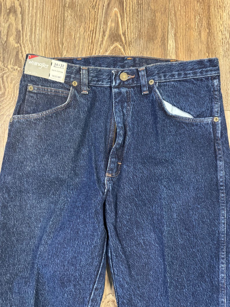 May include: A close-up shot of blue denim Wrangler jeans. The jeans feature a classic five-pocket design, a zip fly, and a button closure. The label indicates a 34x32 size. The denim has a medium wash.