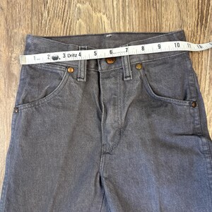 May include: Close-up of gray denim pants with a measuring tape across the waist. The pants have classic five-pocket styling with copper-colored rivets and a button closure. The waist measures approximately 10 inches.