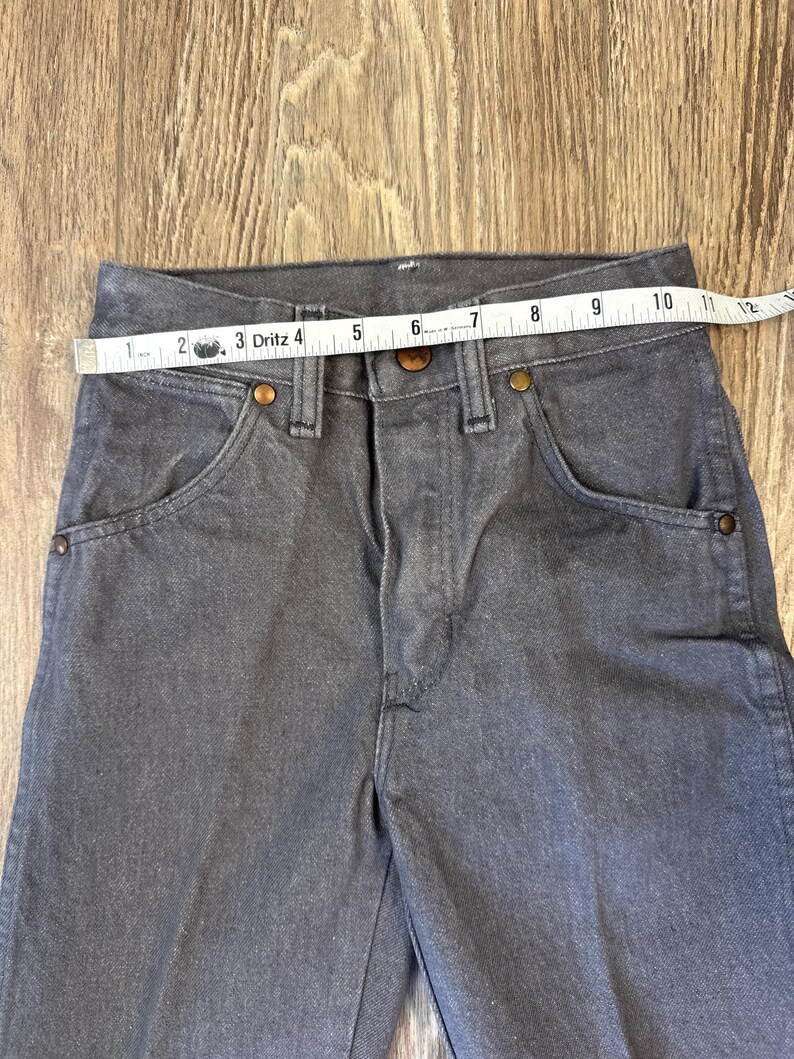 May include: Close-up of gray denim pants with a measuring tape across the waist. The pants have classic five-pocket styling with copper-colored rivets and a button closure. The waist measures approximately 10 inches.