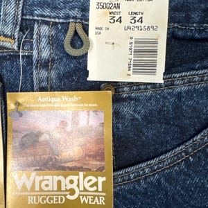Puede incluir: Primer plano de unos vaqueros Wrangler azules con etiquetas. Las etiquetas indican una talla de cintura de 86 cm y una longitud de 86 cm. Los vaqueros est&aacute;n hechos de 100% algod&oacute;n y presentan el estilo "Antique Wash". La etiqueta tambi&eacute;n incluye el texto "Rugged Wear".