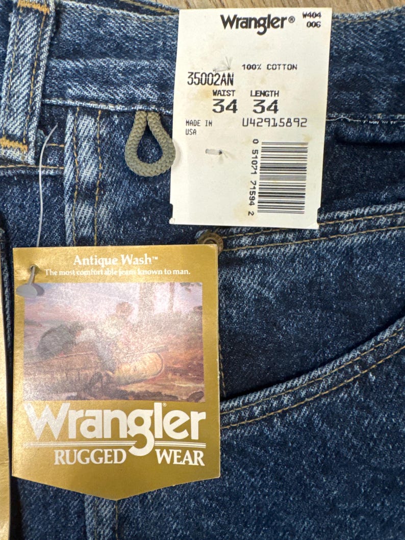 Puede incluir: Primer plano de unos vaqueros Wrangler azules con etiquetas. Las etiquetas indican una talla de cintura de 86 cm y una longitud de 86 cm. Los vaqueros est&aacute;n hechos de 100% algod&oacute;n y presentan el estilo "Antique Wash". La etiqueta tambi&eacute;n incluye el texto "Rugged Wear".